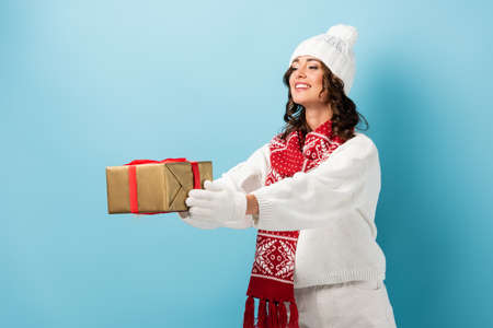 young pleased woman in winter outfit with outstretched hands holding wrapped present on blueの写真素材