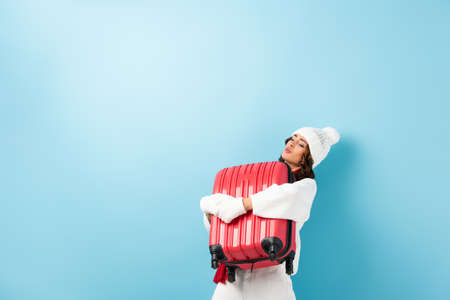 young woman in winter outfit carrying heavy suitcase on blueの写真素材