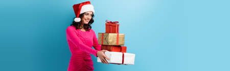 horizontal image of young joyful woman in santa hat and dress holding wrapped presents on blue