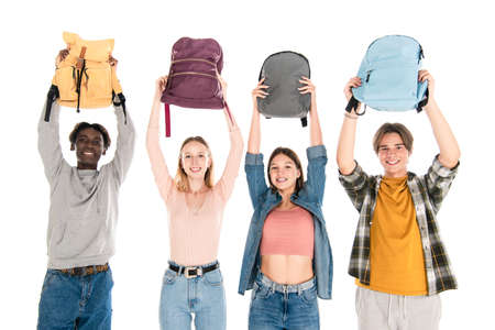 Cheerful multicultural teenagers holding backpacks and looking at camera isolated on white