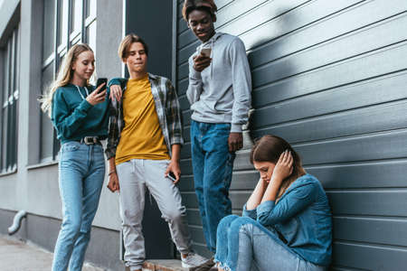 Selective focus of sad girl covering ears near multiethnic teenagers with smartphones and buildingの写真素材