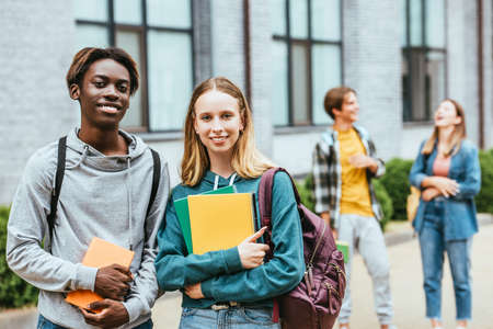 Selective focus of smiling multiethnic teenagers with backpacks and notebooks looking at camera outdoorsの写真素材