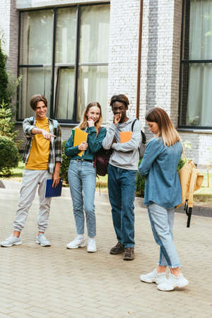 Smiling multiethnic teenagers pointing with finger at sad girl outdoors