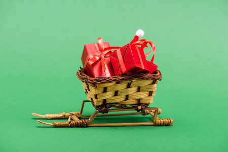 red gift boxes and santa hat in wicker basket on decorative sleigh on green background