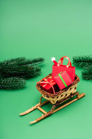 gift boxes and shopping bag with santa hat in wicker basket on decorative sleigh near fir branches on greenの写真素材