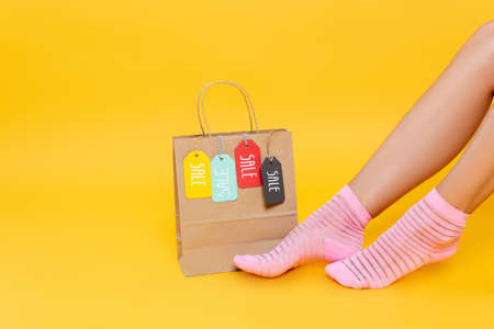 Cropped view of female legs in socks with pink stripes near paper bag with tags with sale words isolated on yellowの写真素材