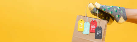 Paper bag with sale tags hanging on woman legs in socks isolated on yellow, banner
