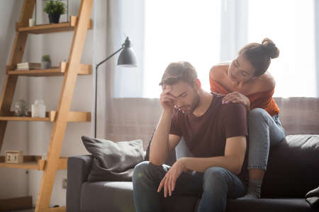 tender woman touching shoulder of worried man sitting on sofa with bowed head and closed eyesの写真素材