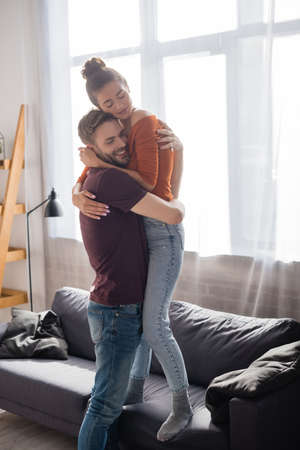 happy man hugging beloved girlfriend standing on sofa at home