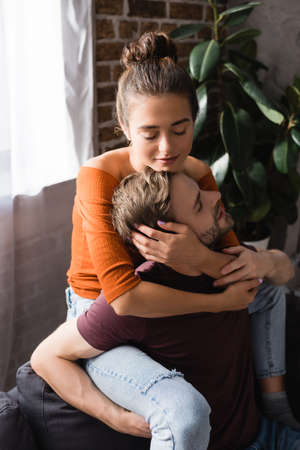 tender woman with closed eyes embracing young boyfriend while sitting on sofa