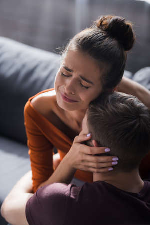 crying woman touching head of beloved boyfriend while sitting on sofaの写真素材