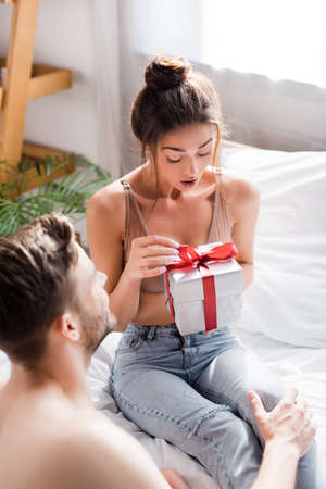 amazed girl in jeans and bra opening gift box near man on blurred foregroundの写真素材