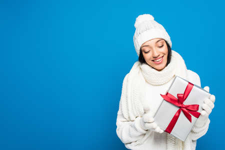 happy woman in winter outfit holding christmas present isolated on blueの写真素材