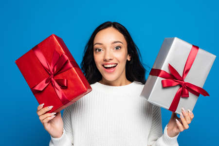 excited young woman in knitted sweater holding gift boxes isolated on blue