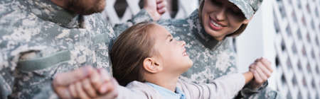 Cheerful girl with outstretched hands sitting near mother and father in camouflages, banner