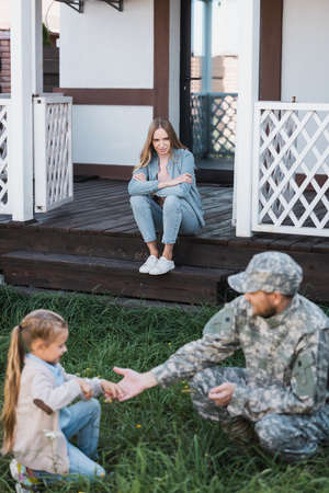 Woman with crossed arms sitting on house threshold on backyard with blurred military man and girl on foregroundの写真素材