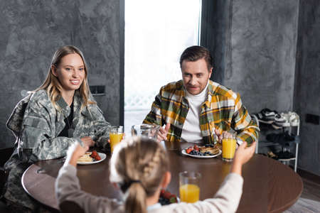Smiling couple having breakfast, while sitting at table in kitchen with blurred girl on foreground