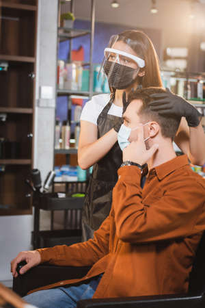 barber in face shield and latex gloves touching hair of client pointing with fingerの写真素材