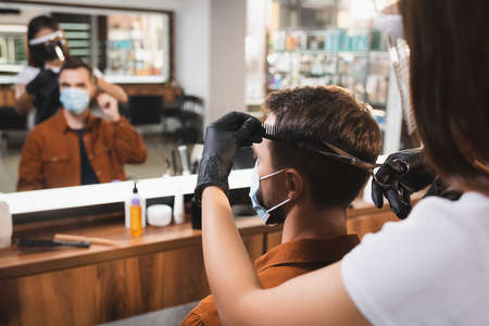 hairdresser in face shield and latex gloves cutting hair of man with blurred mirror reflectionの写真素材