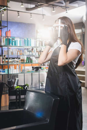 hairstylist in apron fixing face shield while standing near armchair