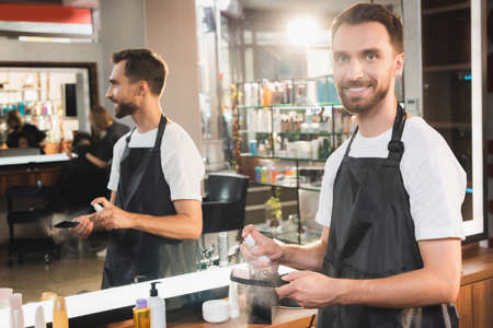 smiling hairdresser disinfecting comb with sanitizer while standing near mirror