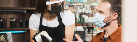 hairdresser in face shield holding container with hair balsam near man in medical mask pointing with finger, bannerの写真素材