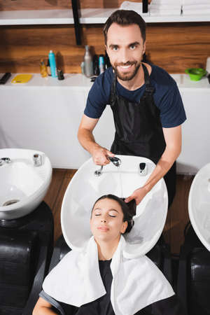 smiling, bearded hairdresser washing hair of young woman in barbershop