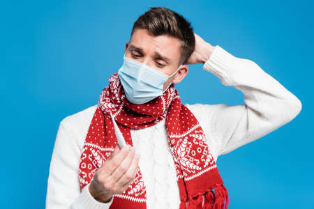 Young adult man with hand near head, wearing medical mask and looking at thermometer isolated on...