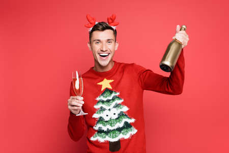 Cheerful man in christmas sweater and headband holding bottle and glass of champagne on red...
