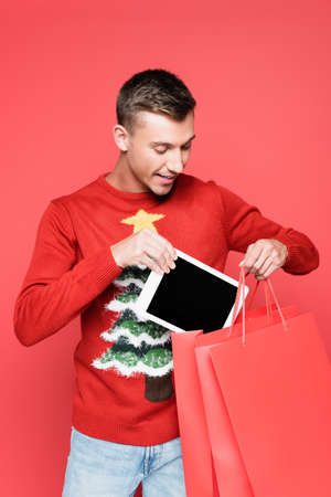 Positive man in sweater with pine tree putting digital tablet in shopping bags isolated on redの写真素材