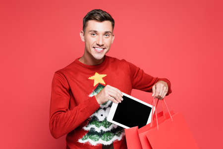 Smiling man in sweater with christmas tree putting digital tablet in shopping bag isolated on redの写真素材