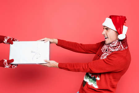 Excited man in santa hat and scarf pulling gift box on red background