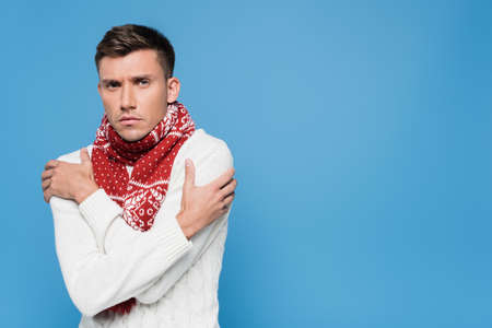 Young man in sweater and scarf feeling cold isolated on blue