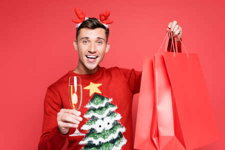 Smiling man in christmas sweater holding glass of champagne and shopping bags isolated on redの写真素材