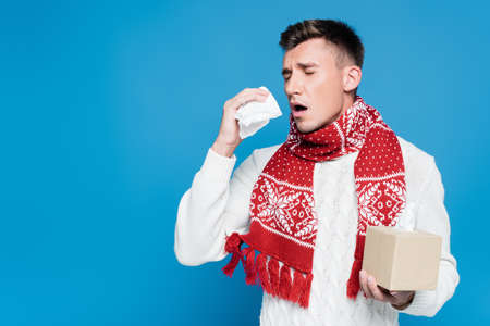 Young adult man in red scarf sneezing, while holding box of tissues isolated on blue