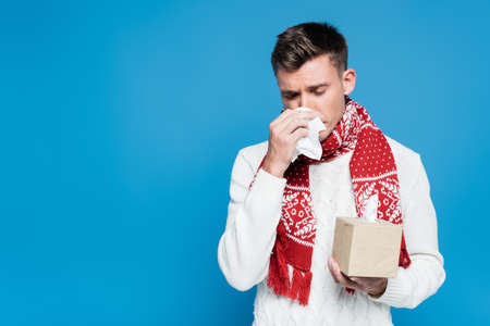 Young adult man in red scarf sneezing, while holding tissue near nose isolated on blue