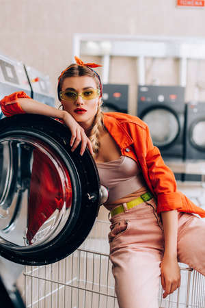 young and stylish woman in headband and sunglasses in modern laundromat