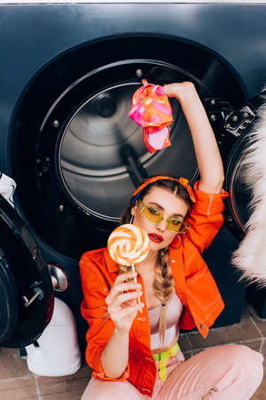 high angle view of fashionable woman holding sweet lollipop and clothing in laundromatの写真素材
