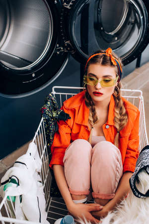 stylish woman sitting in cart with clothing near washing machines in laundromatの写真素材
