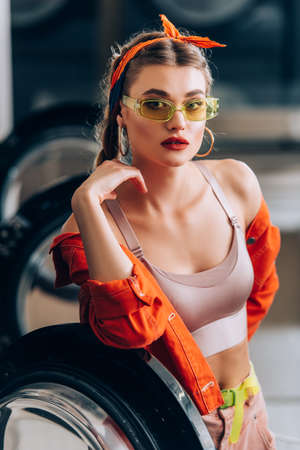 selective focus of trendy woman in sunglasses looking at camera near washing machine