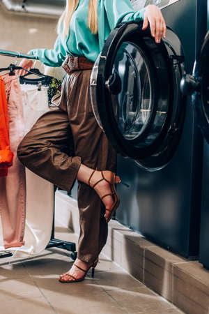 cropped view of stylish woman standing near clothing rack and washing machine in modern laundromat