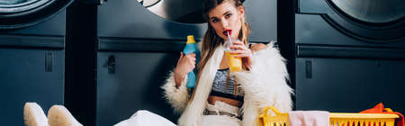 woman in faux fur jacket holding plastic cup with orange juice near basket with laundry in...