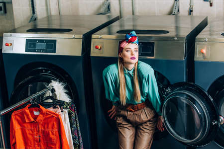 young woman in turban standing near washing machines and clothing rack in laundromat