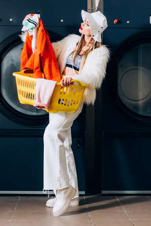 stylish young woman in faux fur jacket and hat holding basket and looking at laundry near washing...