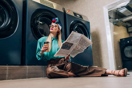 trendy woman in glasses and turban holding paper cup and newspaper while sitting on floor in laundromatの写真素材