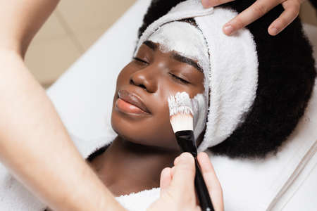Close up view of man applying face mask with cosmetic brush on cheek of african american woman in...
