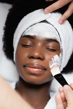 Top view of african american woman lying, while spa therapist applying face mask on cheek with cosmetic brush in spa salonの写真素材