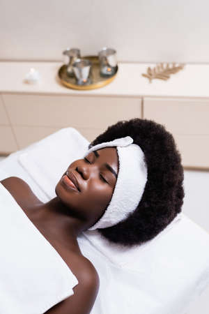 African american woman in white headband covered with bed sheet lying on massage table in spa salon on blurred backgroundの写真素材