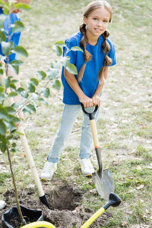 girl looking at camera while holding shovel near mother and young green tree, ecology concept