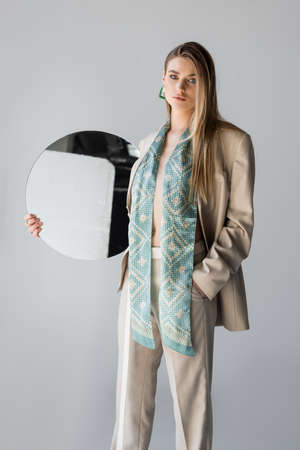 young woman in suit holding round mirror and standing with hand in pocket on grayの写真素材